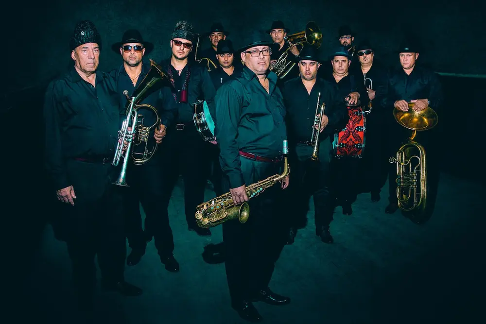 Fanfare Ciocărlia – muzycy orkiestry z instrumentami dętymi na ciemnym tle